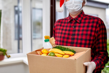 Volunteer man in Santa hat and protective mask and gloves delivery donation box at home in Christmas
