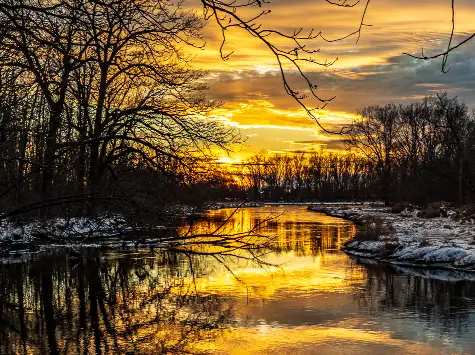 Sunset over a calm river with snow-covered banks and leafless trees.