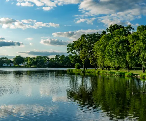 Calm lake bordered by lush green trees under a partly cloudy blue sky.