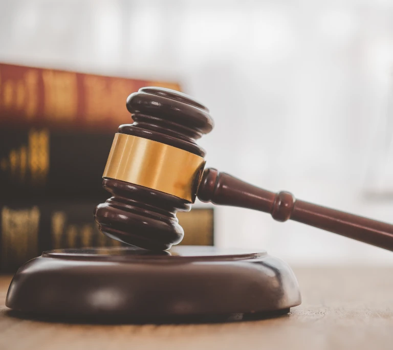 Wooden judge's gavel with gold band resting on sound block, blurred legal books in background.