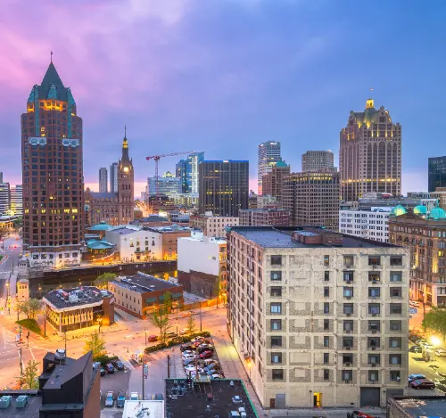Dusk cityscape featuring illuminated historic and modern buildings under a purple-blue sky.
