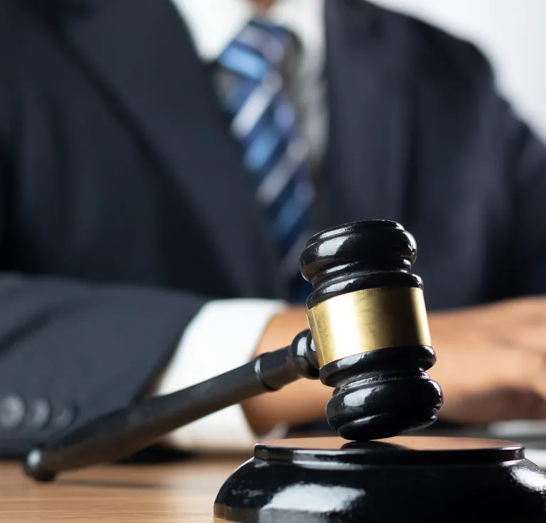 Close-up of a black judge’s gavel with a gold band on a wooden desk, person in suit blurred background.