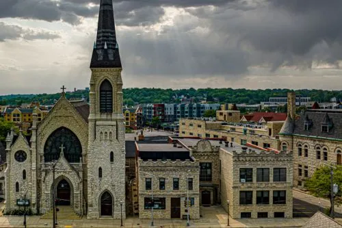 A historic stone church with a tall steeple amid urban buildings under a cloudy sky.