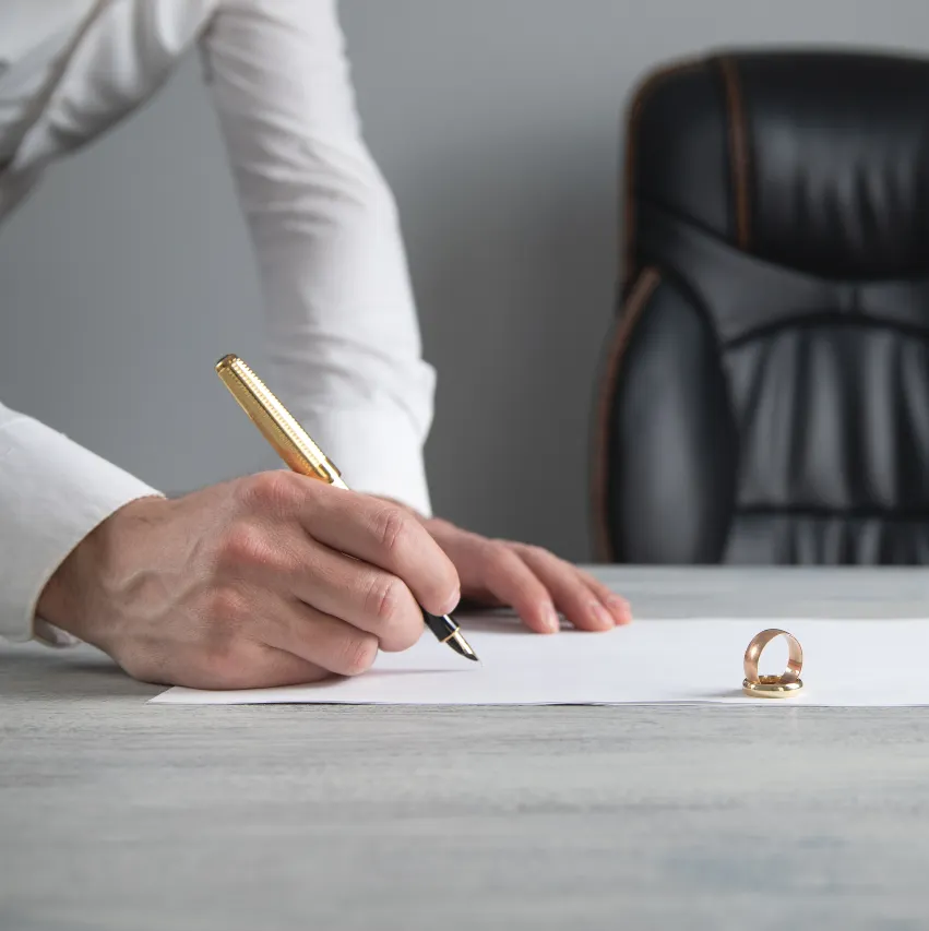Person in white shirt signing document with gold pen, two rings on desk nearby.