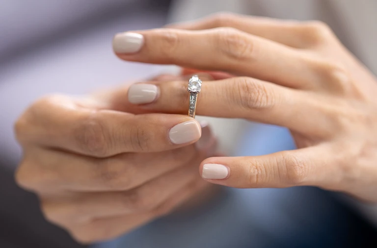 Close-up of hands with light-colored polished nails holding a silver engagement ring with a round diamond.