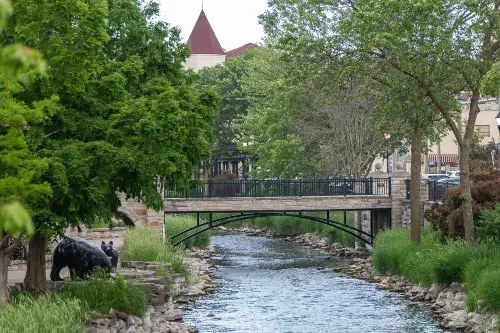 View of a small stone-lined river with a pedestrian bridge and surrounding green trees.
