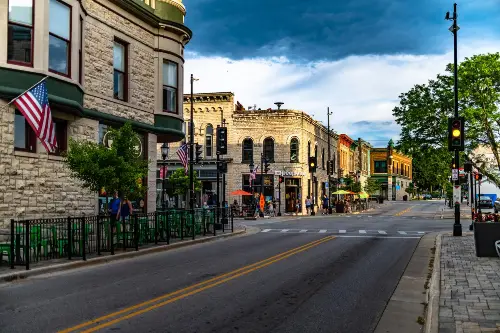 Urban street scene with historic buildings, American flags, pedestrians, and traffic lights under a cloudy sky.