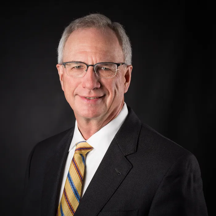 Professional portrait of an older man wearing glasses, a dark suit, white shirt, and striped yellow tie.