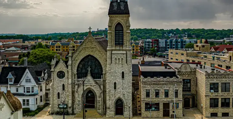 Stone Gothic-style church with a tall steeple amidst urban buildings under cloudy sky.