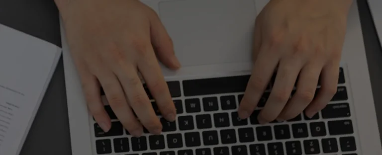 Close-up of hands typing on a black and silver laptop keyboard with a document nearby.