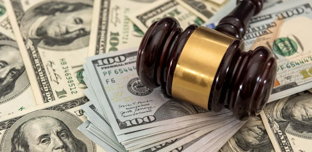 Dark wooden judge's gavel with brass band resting on a stack of US hundred-dollar bills.