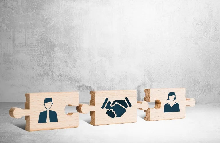 Three connected wooden puzzle pieces on a gray surface, featuring icons of a man, a handshake, and a woman.