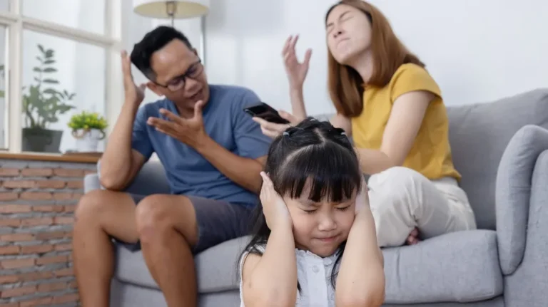 Young girl covering ears in distress while parents argue on couch behind her.