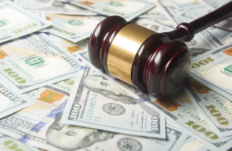 Wooden judge's gavel with a gold band resting on scattered hundred-dollar bills.
