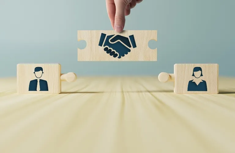 Hand holding wooden puzzle piece with handshake icon between male and female avatar puzzle pieces.