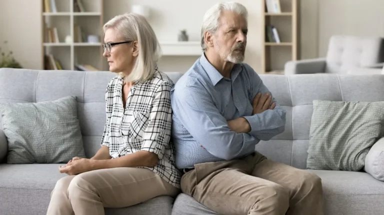 Elderly man and woman sitting back-to-back on gray sofa, both looking away with serious expressions.